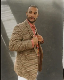 Man wearing a brown plaid blazer and a pink shirt standing against a dark grey textured studio backdrop.