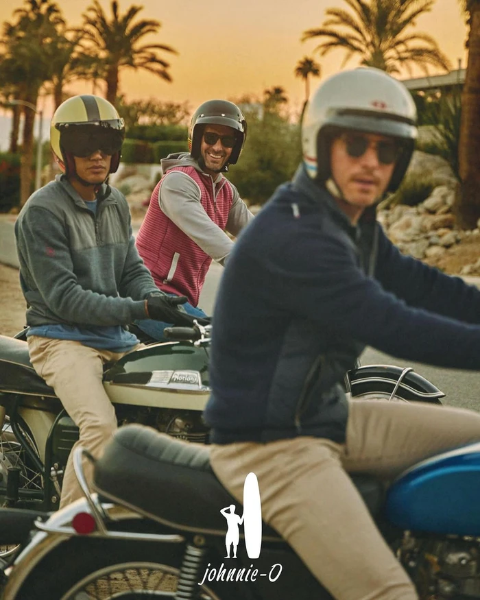 Three men wearing helmets and sunglasses riding vintage motorcycles along an outdoor street at sunset.