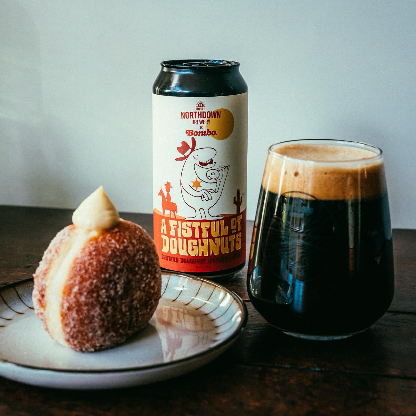 Craft stout in glass, donut with cream, and labeled beer can featuring cartoon sheriff on a brown wooden table indoors.