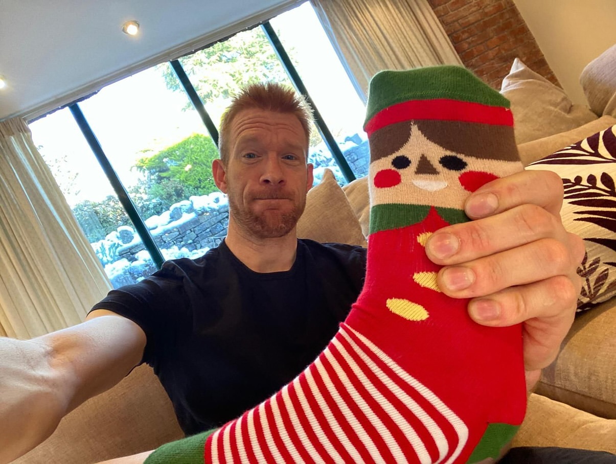 Adult man takes a selfie indoors, holding up a sock with a cartoon elf design, in front of a window with greenery visible outside.
