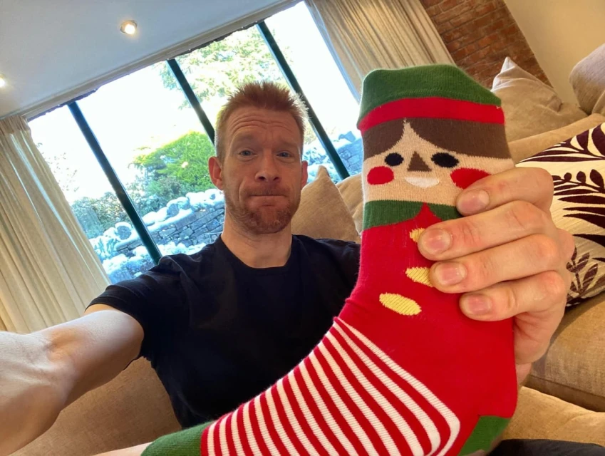 Adult man takes a selfie indoors, holding up a sock with a cartoon elf design, in front of a window with greenery visible outside.