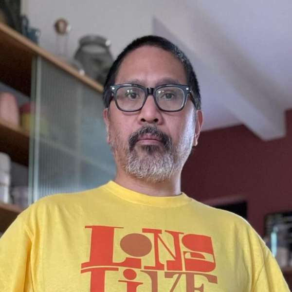 Adult male in eyeglasses and yellow graphic tee poses indoors against a bookcase. Head and shoulders shot; neutral facial expression.