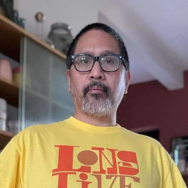 Adult male in eyeglasses and yellow graphic tee poses indoors against a bookcase. Head and shoulders shot; neutral facial expression.