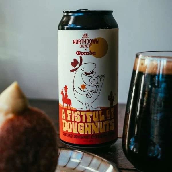 Beer can featuring cartoon cowboy with a donut, a glass of dark beer, and a donut on a wooden surface, product shot.