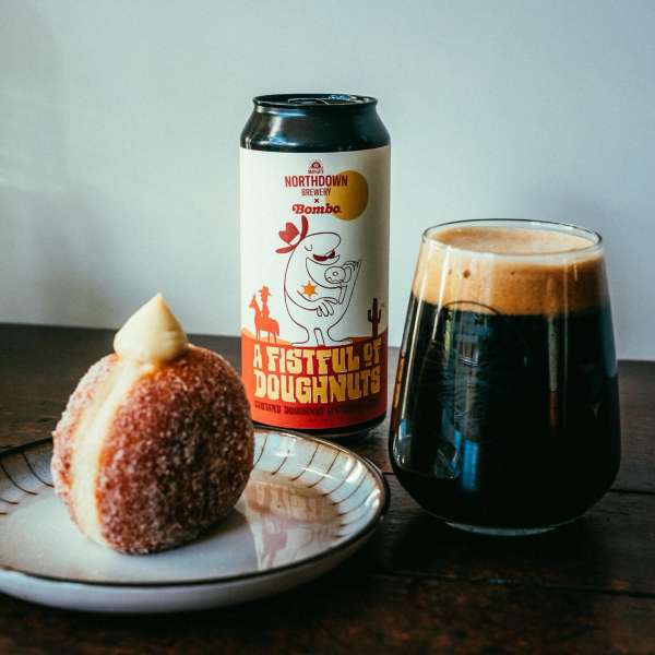 Craft beer and sugar-coated donut on a plate. The can shows cowboy cartoon art and says 'A Fistful of Doughnuts' on a wood surface.
