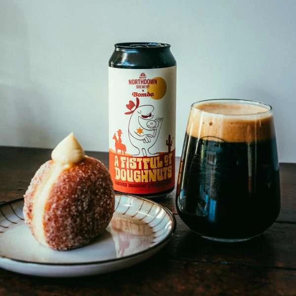 Craft beer and sugar-coated donut on a plate. The can shows cowboy cartoon art and says 'A Fistful of Doughnuts' on a wood surface.