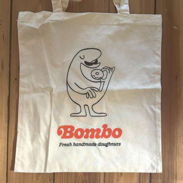 White tote bag with cartoon graphic of a character holding a donut and the text "Bombo, Fresh handmade doughnuts."