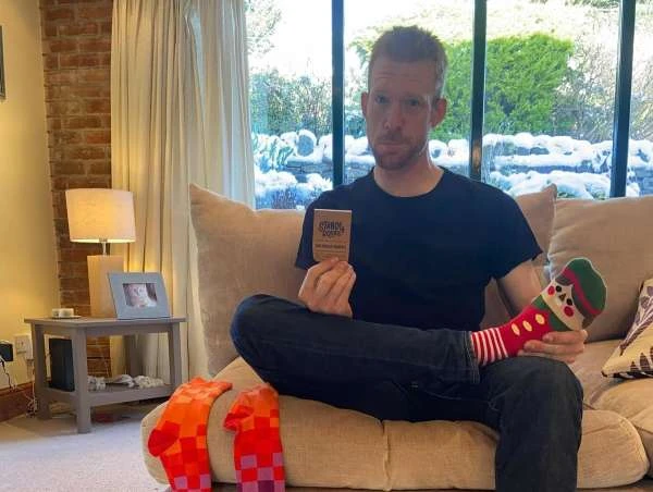 A man in a black shirt sits on a couch holding a sock product box, with colorful socks displayed, inside a cozy home setting.