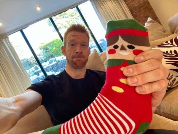 Adult man holding a red and green Christmas elf sock in a casual indoor setting with a view through a large window.