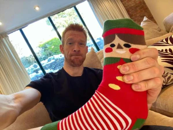 Adult man holding a red and green Christmas elf sock in a casual indoor setting with a view through a large window.
