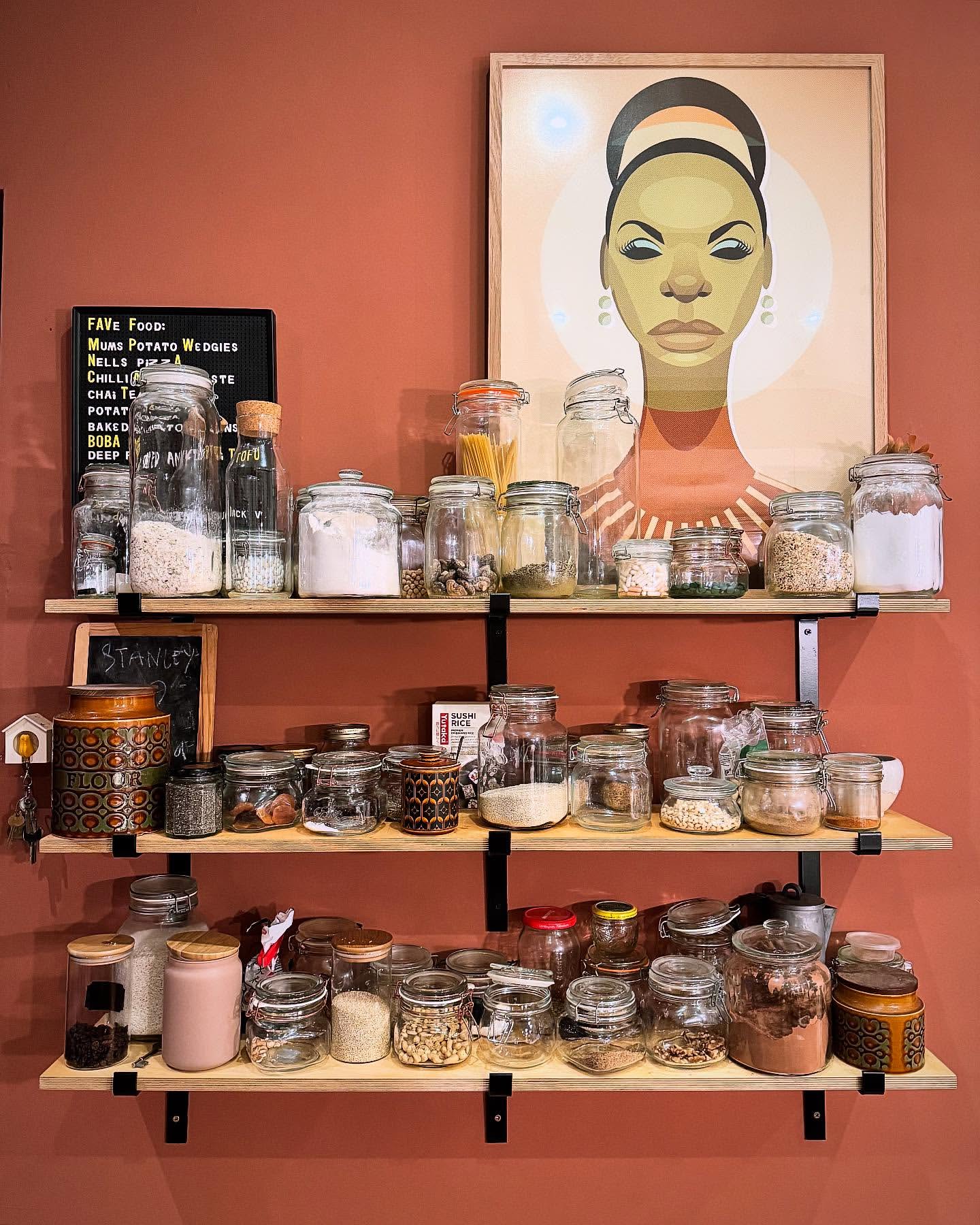 Pantry shelves with glass jars filled with food items, plus an illustration of singer Nina Simone on the top shelf, against a brick-red wall.