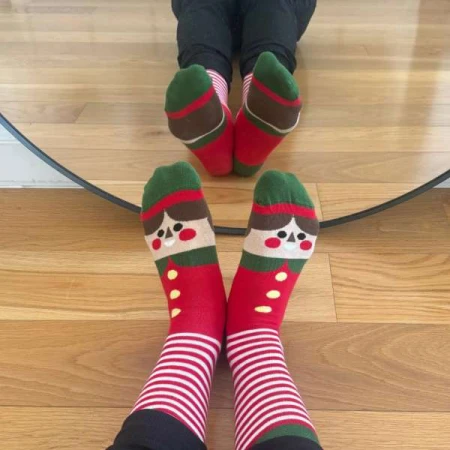 Festive holiday elf socks on feet, featuring red, green, and white stripes with cartoon elf face on light-wood floor in a cozy setting.