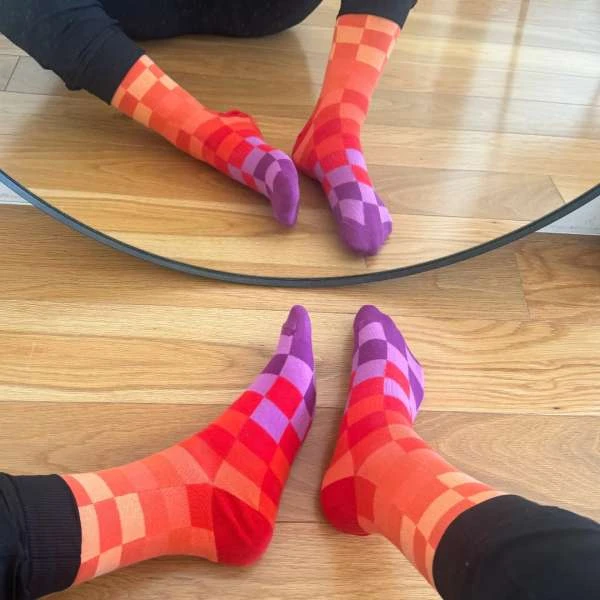 Close-up of two pairs of legs wearing checkered, colorful socks reflected in a mirror on a light-colored wooden floor, indoor setting.