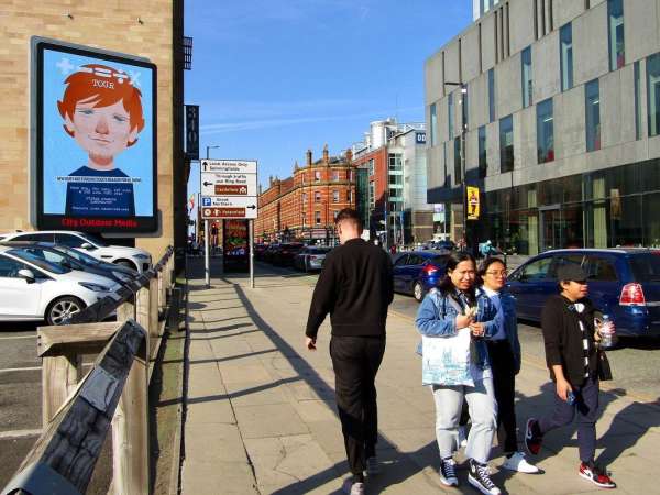Outdoor advertising billboard displays an animated figure as pedestrians walk on a city sidewalk during the daytime.