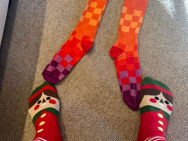 Close-up on feet wearing novelty socks, including a red and green elf sock and an orange checkerboard pattern sock.