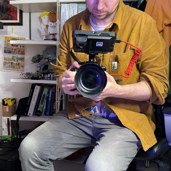 Man holding a professional video camera setup with monitor and large lens in an indoor environment with shelves in the background.
