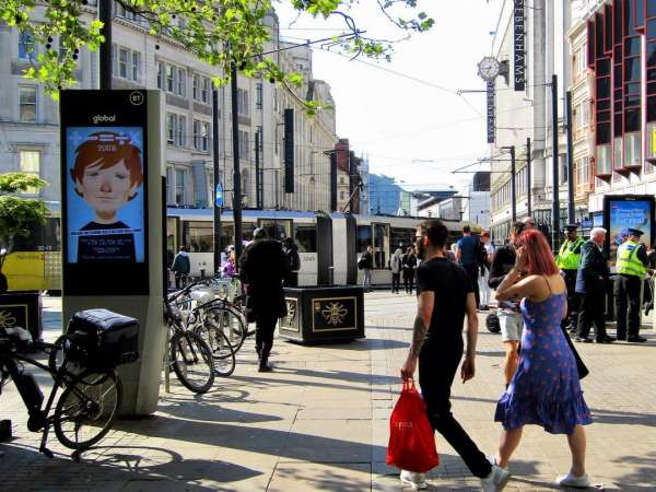 People walk along a city sidewalk with a tram in the background and an advertisement for a cartoon on a digital kiosk.