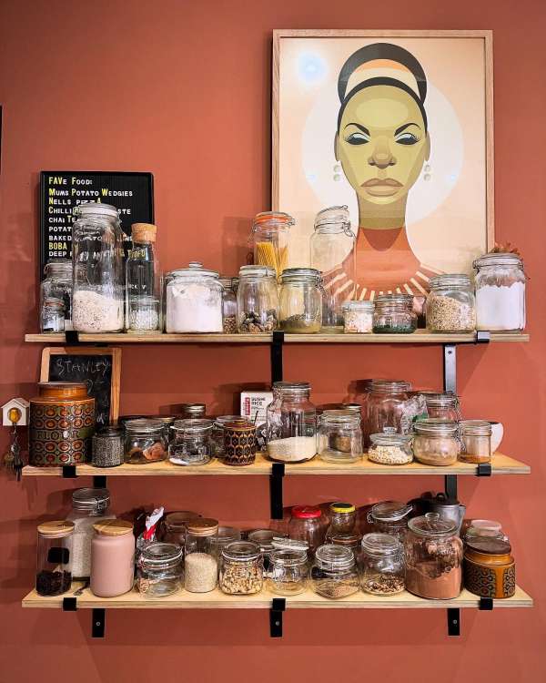 Shelves filled with assorted food in glass jars, set against a reddish-brown wall with framed portrait art hanging on the wall.