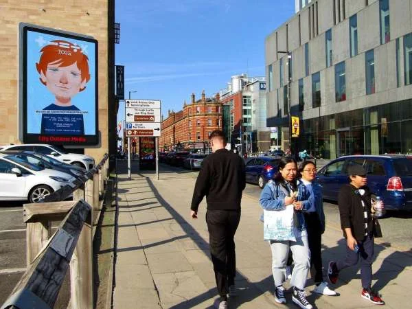 Pedestrians walking along a sunny city sidewalk past parked cars and a large outdoor digital billboard illustration by Stan Chow.