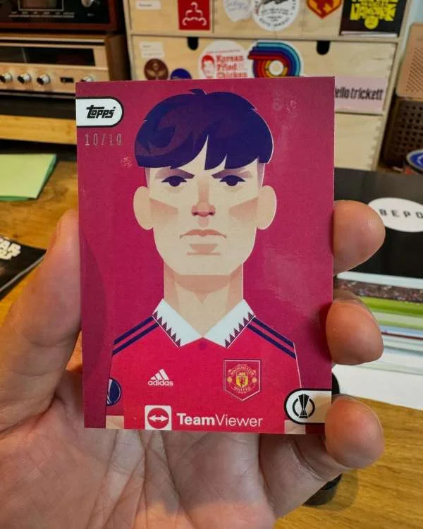 A hand holding a trading card featuring an illustrated portrait of a male soccer player in a red jersey against a pink background.