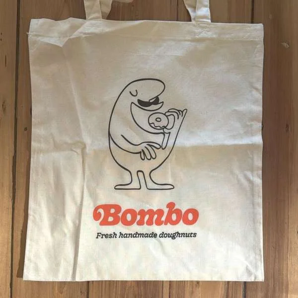 Cream canvas tote bag with black cartoon character illustration eating a doughnut and orange Bombo logo.