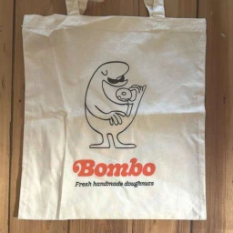 Cream canvas tote bag with black cartoon character illustration eating a doughnut and orange Bombo logo.