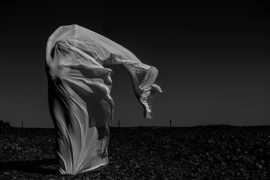 Figure draped completely in white fabric billowing in the wind against a dark horizon and rocky ground.