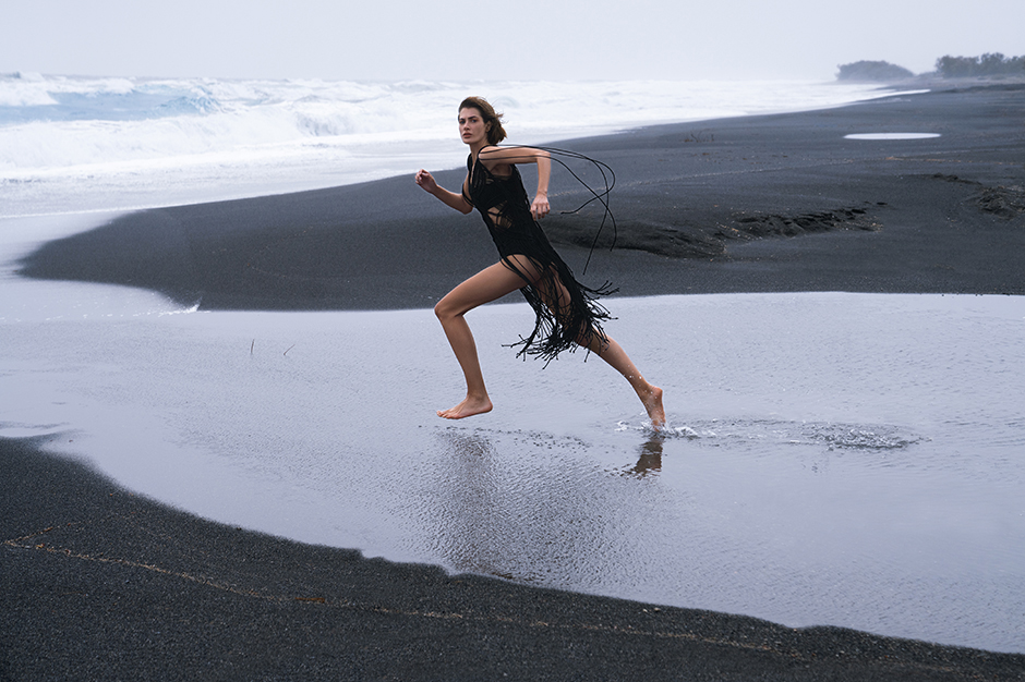 Woman in black fringed dress running barefoot across a dark sand beach with ocean waves