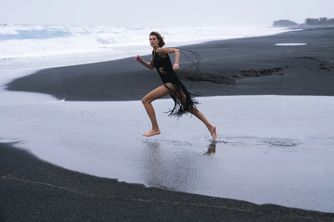 Woman in black fringed dress running barefoot across a dark sand beach with ocean waves