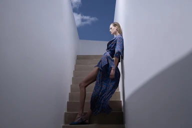 Model in patterned blue wrap dress leaning against a white stairwell wall under a patch of blue sky styled by Sandy Karagianni