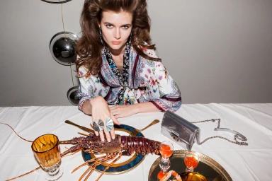 Model with voluminous brown hair wearing a patterned top seated at a white table with a lobster, silver handbag, and glassware.