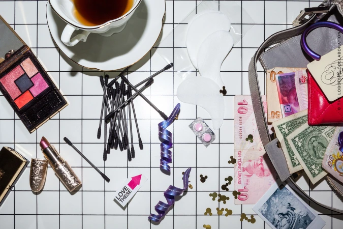 Flat lay of makeup, teacup, cotton swabs, currency, and accessories on a black and white grid background styled by Sandy Karagianni.