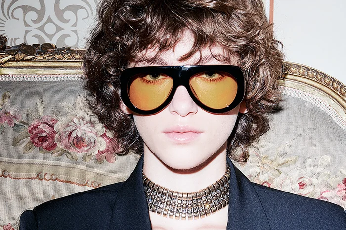 Model with curly brown hair wearing black sunglasses with orange tinted lenses and a gold choker necklace.
