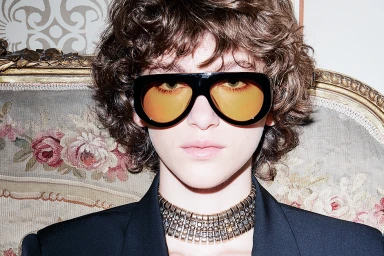 Model with curly brown hair wearing black sunglasses with orange tinted lenses and a gold choker necklace.