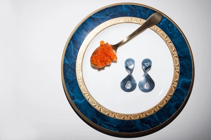 Caviar on a spoon and two earrings resting on a white and blue decorative plate against a white background