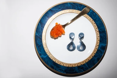 Caviar on a spoon and two earrings resting on a white and blue decorative plate against a white background