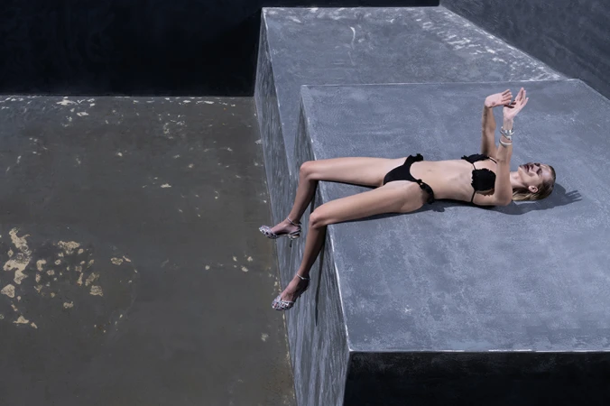 Model wearing a black bikini and silver heels reclining on a large grey concrete geometric structure.
