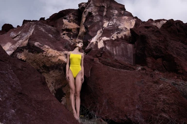 Model in a bright yellow one-piece swimsuit standing against towering dark volcanic rock formations.