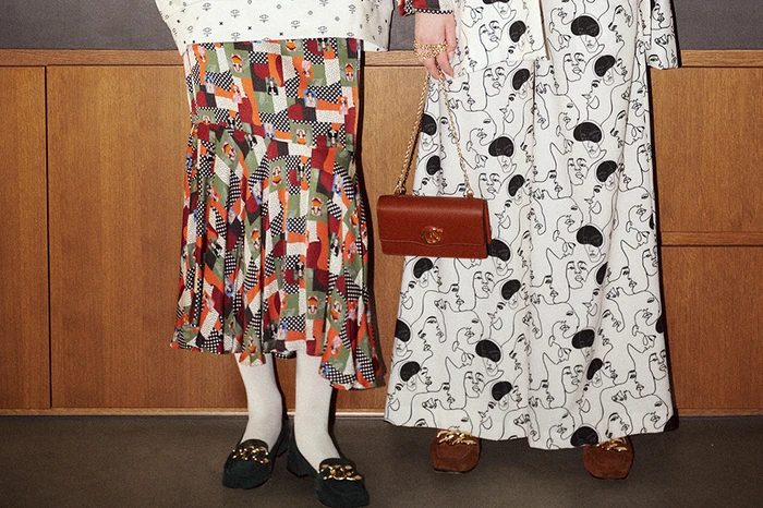 Two people wearing patterned outfits and loafers, one holding a brown leather shoulder bag against wood paneling.