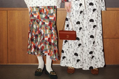 Two people wearing patterned outfits and loafers, one holding a brown leather shoulder bag against wood paneling.