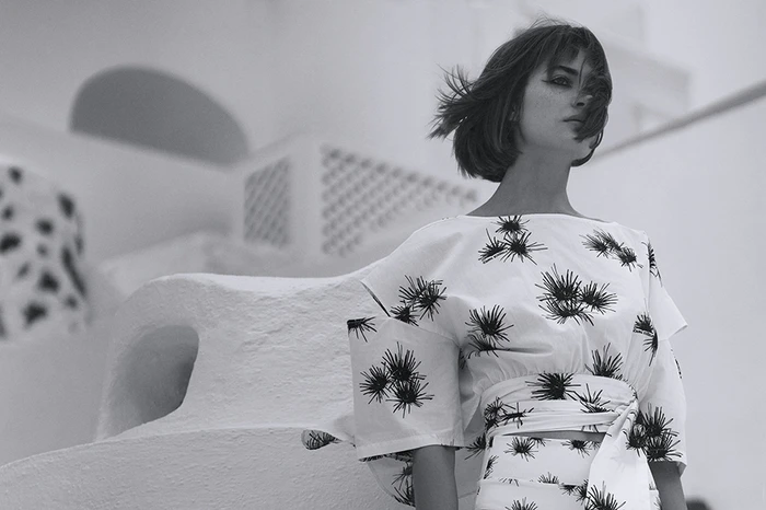 Black and white portrait of a woman with short hair wearing a patterned white top against an outdoor architectural background.
