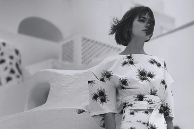 Black and white portrait of a woman with short hair wearing a patterned white top against an outdoor architectural background.