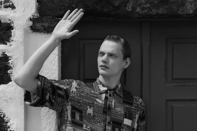 Monochrome portrait of a man wearing a patterned shirt with his right arm raised to shade his face against a rustic stone and wood wall.