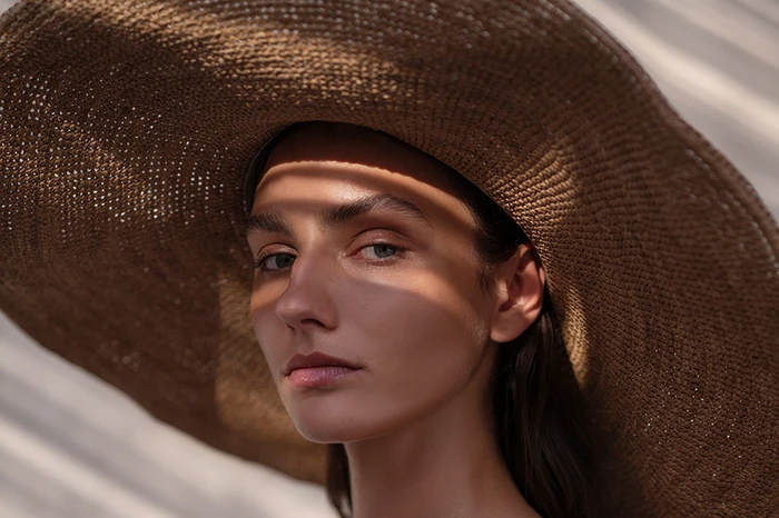 Close-up portrait of a woman wearing a large brown woven sun hat with striking diagonal shadows cast across her face.