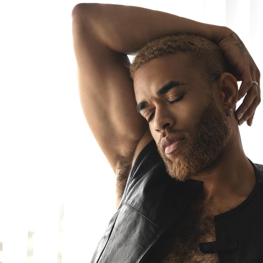 Portrait of a man with blonde hair and beard in a black vest, eyes closed, arm raised, and wearing hoop earrings against a bright background.