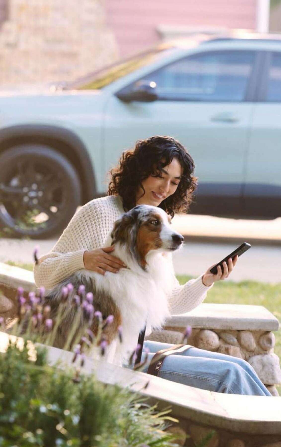 Woman with curly hair sits with an Australian Shepherd on a stone bench outdoors, holding a cell phone, with a light green car in the background.