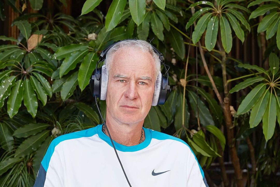 Portrait of adult male wearing headphones, outside, surrounded by leafy green plants; the subject is wearing a white and blue shirt.