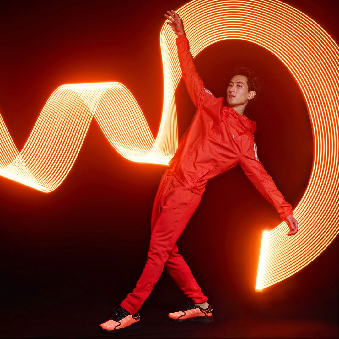 Athletic male in red tracksuit strikes dynamic pose in front of an orange light trail against a dark background, creating a striking contrast.