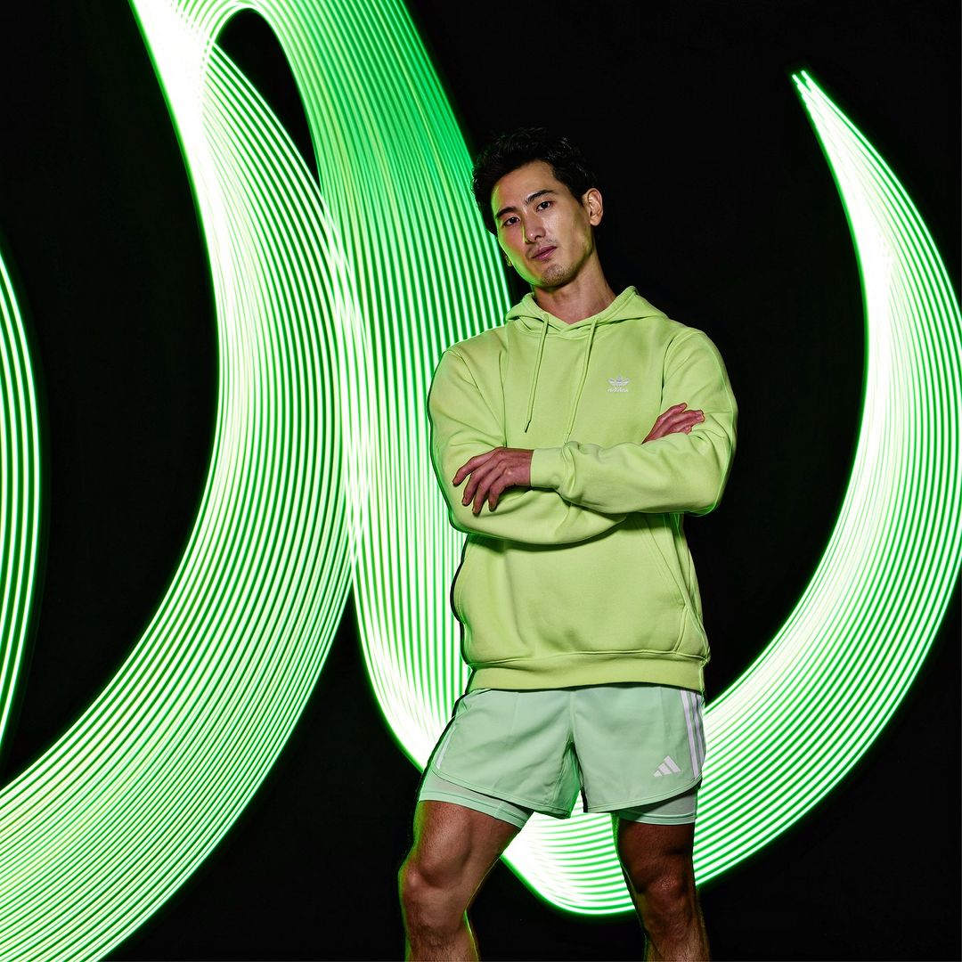 Man in lime green hoodie and shorts standing with arms crossed against a black background with looping green light trails.