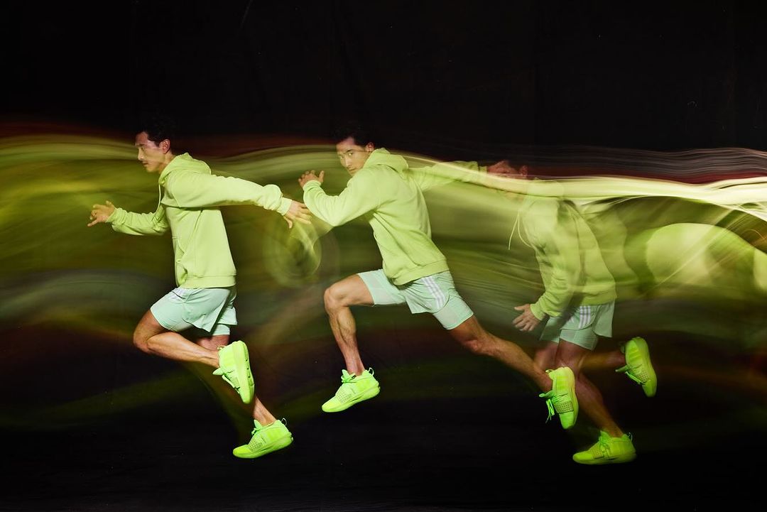 Man in motion wearing a green hooded sweatshirt and shorts running against a black background, captured with a long exposure technique.